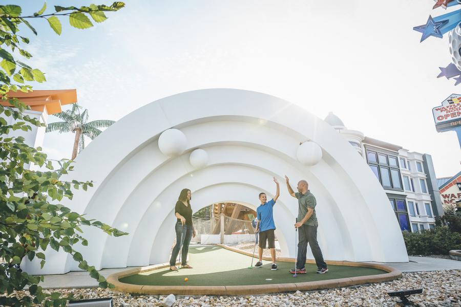 Family of three high five while playing mini golf in front of large white arch