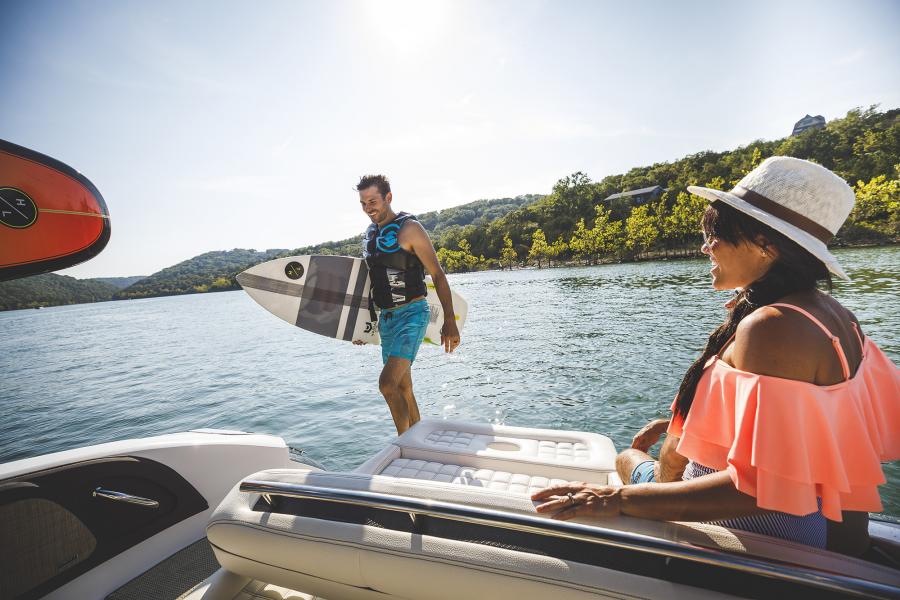 Man walks up from back of a boat with wakeboard in hand as woman looks on