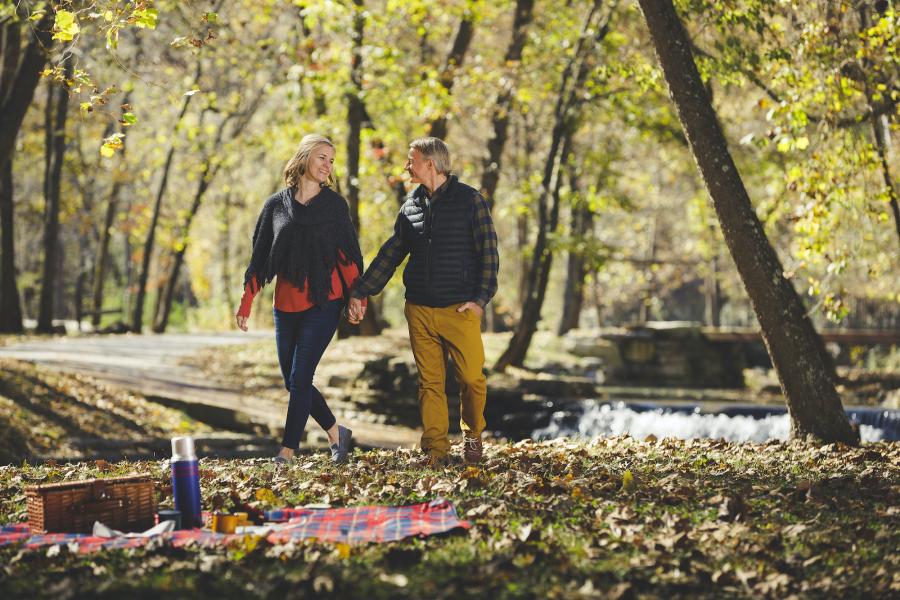 Man and woman holding hands and walking towards a picnic blanket laid out over leaves that have fallen from surrounding trees in Dogwood Canyon.