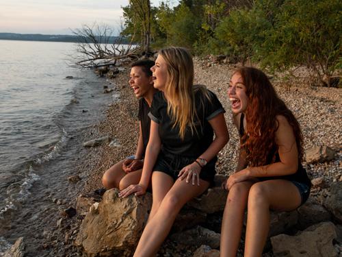 Three girls sitting on a lake shoreline on rocks while staring into the sunset and laughing in Branson.